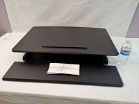 Standing desk riser converter in lowered position with instruction manual on lower shelf and water bottle for scale.