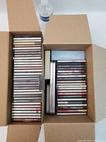 Top view of two open cardboard boxes filled with multiple stacks of compact discs in jewel cases, showing spines with titles visible in some cases.