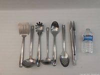 All 8 metal cooking utensils laid out flat with a water bottle for size reference.