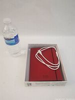 Kobo e-reader covered in red case with white USB charging cable on top, next to a water bottle for scale. Shown inside the box.