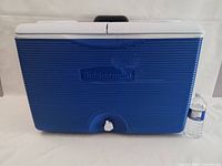 Front view of Rubbermaid rolling cooler showing blue textured body, white dual lid, and front drain valve with water bottle for scale.