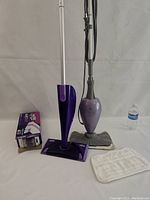 Shark floor steamer in gray and purple with two fabric pads and Swiffer Wet Jet with box of pads side by side.