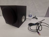 Yamaha black YST-SW012 subwoofer speaker with fabric grille on front and side port, next to water bottle for size reference and cables on white surface.