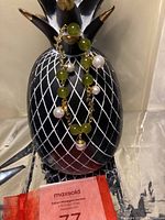 Front view of the charm bracelet draped over a black and white ceramic pineapple display stand, showing the alternating olive green agate stones and white pearls on gold-tone chain.