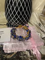 Front angle of handmade blue sapphire and crystal bead copper wire bangle with adjustable hook, placed on pink fabric next to decorative black and white patterned vase.