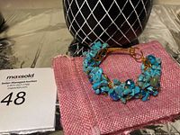 Full view of handmade copper wire bangle with turquoise chip beads and blue faceted crystal beads resting on pink cloth with a black vase behind.