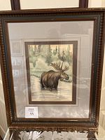 Full view of framed and matted painting showing a moose standing in water with forest in background. The dark brown wooden frame is worn and marked.