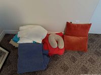 Photo of stacked blankets including blue wool blanket, white/cream and turquoise blankets, two rusty orange throw pillows, and a grey travel neck pillow.