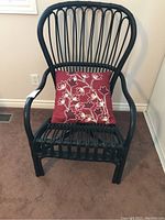 Full view of the black rattan armchair with red floral accent pillow placed on the seat. Chair shows curved armrests and vertical rattan slats on the backrest.