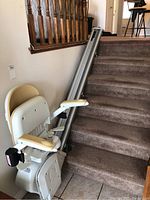 Image of white stair lift chair on carpeted stairs with metal rail. Chair has padded armrests and seat.
