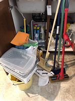 Photo of cleaning supplies under sink showing mops, bucket, glove, plastic container, dish rack, and various cleaning products.