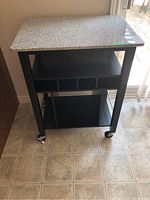 Front view of the black coffee cart showing the stone countertop, lower shelves, and wheels on a tiled floor.
