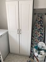 White cupboard with two paneled doors closed, showing silver handles, next to a freezer and ironing board in laundry area.