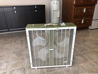 Full view of retro white Lasko box fan on a tiled floor, showing front grille and handle on top.