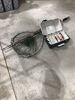 Photo of two fishing poles resting on a large fishing net with a plastic tackle box open showing various lures inside