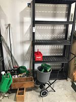 Full view of lot showing black shelving unit, wooden toolbox, two green watering cans, long-handled shovels and forks, gray plastic seeder/spreader, and assorted hand tools including clippers.
