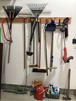 Full view of gardening tools hanging on wooden rack: rakes, bow saw, hoe, broom, dustpan, red gas can, hedge trimmer, gloves and small tools.