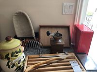 Photo of wooden coffee grinder, biscotti cookie jar, bread board, metal toast rack, woven tray, and red metal tin box arranged on counter