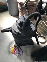 Full view of the black Shop Vac showing the cylindrical body, attached hose, and the filter at base with some debris around.