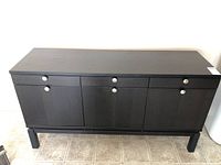 Front view of black wood-grain sideboard showing 3 drawers across top and 3 cupboard doors below with silver round handles, standing on tile floor.