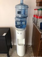 Full view of white floor standing water dispenser with a blue 5-gallon water bottle mounted on top and a second blue 5-gallon bottle placed nearby.