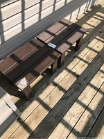 Two outdoor wooden side tables with dark brown stain shown side by side on a wooden deck with sunlight and shadows. The image emphasizes the shape, size, and color of the tables.