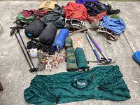 Photo showing multiple hiking backpacks, sleeping bags, camping stakes, hiking poles, and a folded green hammock tent on grey carpeted floor.