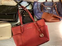 Photo of a large red Michael Kors tote bag, a navy and brown handbag, and various smaller bags arranged on a table