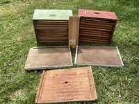 Two wooden beekeeping hive boxes, one green and one red, displayed outside on grass with wooden frames removed and laid in front.