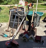 Photo showing the full assortment of garden supplies including a black compost bin, a green wheelbarrow, various garden tools, bird houses, and a Stihl weed whacker on concrete surface.
