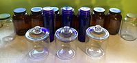 All jars grouped with dark brown gallon jars in back left, tall blue glass jars in center back, clear gallon jars on far left and far right, and three large clear lidded jars in front row