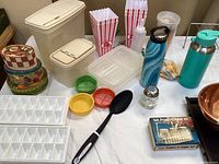 Overview of assorted kitchen items including containers, popcorn boxes, sprouting lids, water bottles, utensils and copper bowls on a table.
