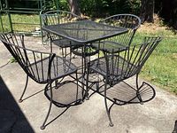 Full view of outdoor black metal patio table with four matching chairs placed on concrete patio showing general wear and paint loss spots.