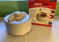 Nesco Snackmaster Entrée Food Dehydrator with box, showing front and top view of the white appliance with label on top and stacked round trays.