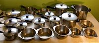 Wide view of the complete lot laid out on a table showing a variety of sizes and styles of RevereWare stainless steel pots, pans, and bowls, mostly with lids featuring black knob handles. Some lids and handles show slight wear.