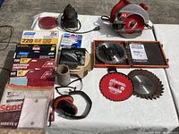 Skilsaw circular saw, black palm sander, multiple sandpaper sheets in various grits, extra blades, safety glasses, earmuffs, and AvantiPro 8-inch stacked dado blade kit in wooden box with manual all laid out on table.