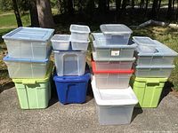 Photo showing a collection of various plastic storage bins stacked and arranged outdoors on a paved area with trees and grass in background.