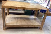 Front view of sturdy wooden workbench with attached blue metal vice on right side and removable wooden side tray next to it. Solid lower shelf for storage.