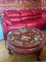 Full view of red round coffee table with glass top placed in front of a red leather sofa, showing floral gold decor on the top and sides.