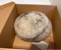 Top and angled views of a round fox fur hat placed inside a cardboard box. The hat shows natural variegated colors of white, gray, and brown fur with a dense and plush texture.