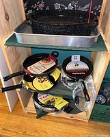 Photo of shelves showing large speckled enamel roasting pan and metal sheet roasting pan on top shelf, various frying pans with packaging on middle and lower shelves, and a speckled enamel wok.