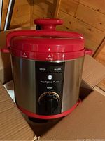Wolfgang Puck pressure cooker inside a cardboard box, showing the entire unit from front and top angle, highlighting stainless steel body and red lid with handle.