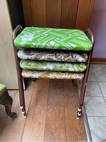 Four retro stools stacked showing metal frames and four distinctly upholstered seats: one green and white geometric pattern, three paisley patterns in brown, green, and gold tones. Close-up view of upholstery and stool build.