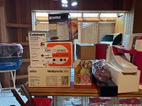 Wide view of kitchen counter showing boxed Kenmore coffee grinder, Cuisinart parts, Braun hand blender set, Starfrit hand juicer, and some packaged food products and accessories.