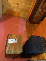 Photo showing two speakers, one wood grain finish and one black finish, placed on the floor in basement.