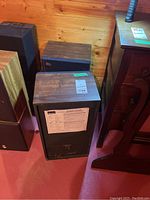 Two wooden-cabinet speakers with black fabric front grills placed on basement floor near wooden panel wall. Label on back of each speaker visible.