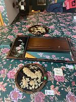 Two Asian style wood wall art pieces displayed on floral wallpaper including a rectangular mirror with decorative floral and butterfly inlays and a round carved wood plaque featuring a bird and flowers.