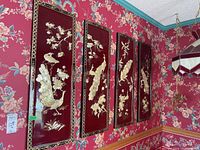 Four vertical rectangular wall art panels with red backgrounds, mother of pearl peacock and flower designs, black and gold borders, mounted on floral wallpapered wall.