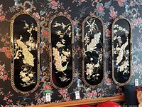 Four oblong black wall art panels with Asian-inspired bird and floral designs hung side-by-side on a decorative floral wallpapered wall.