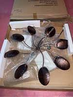 Full top view of the copper kinetic windmill showing eight curved copper blades attached to thin copper arms radiating from the center, sitting in protective foam and bubble wrap inside the box.